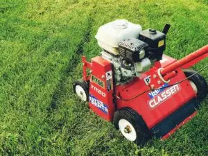 A red Classen TR-20 walk-behind dethatcher with a white fuel tank and black engine, sitting on vibrant green grass. The machine has four wheels with white hubs and black tires, and a red handle extends towards the right. "P766" and "TATES RENTS" are visible on the left side, while "CLASSEN" branding is on the right.