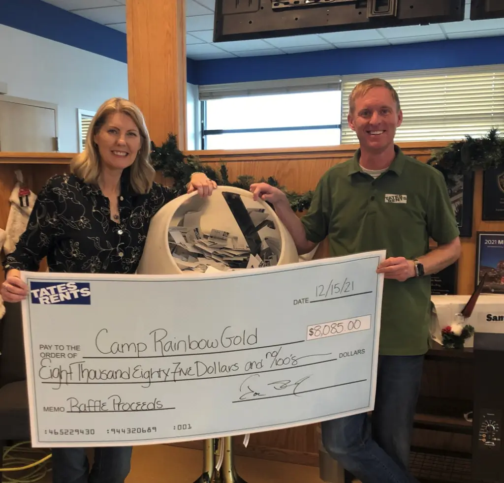 A woman and a man stand side-by-side, holding a large check from "Tates Rents" made out to "Camp Rainbow Gold" for $8,085.00. The check is dated 12/15/21 and has "Raffle Proceeds" in the memo line. The woman on the left wears a dark patterned shirt, and the man on the right wears a green polo shirt with a "Tates Rents" logo. A white raffle drum filled with tickets is visible between them.
