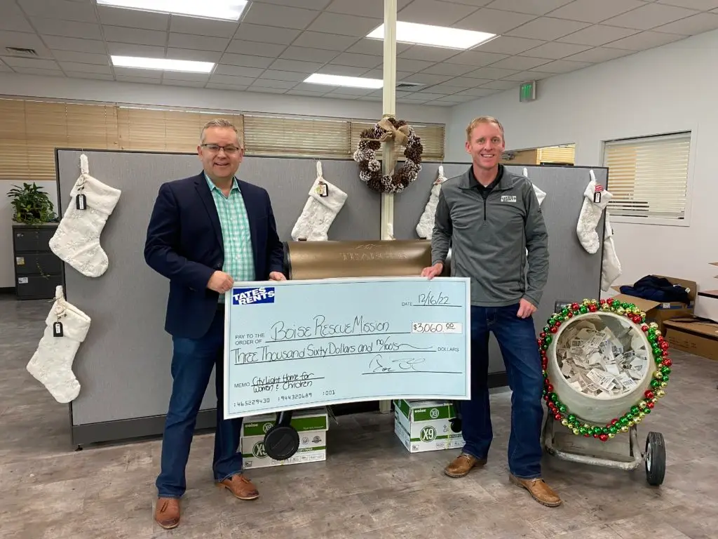 Two men, one in a blue blazer and one in a gray polo, stand in an office holding a large check for $3,060 made out to Boise Rescue Mission. Behind them are office cubicles decorated with white Christmas stockings, a pinecone wreath, and a concrete mixer with Christmas ornaments and slips of paper inside.