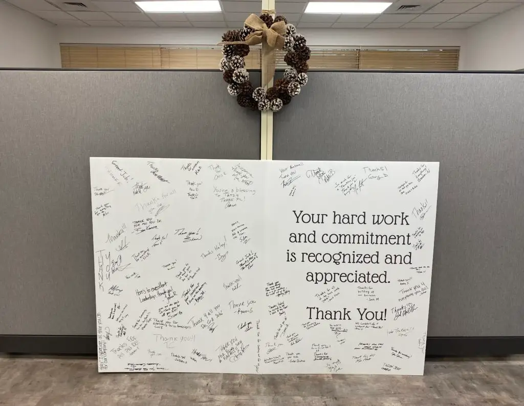 A large white board filled with numerous handwritten thank you messages and signatures, displayed in an office cubicle setting. A central message in black reads, "Your hard work and commitment is recognized and appreciated. Thank You!" A decorative pinecone wreath with a burlap bow hangs above the board on the cubicle partition. The background shows grey cubicle walls, light brown blinds, and a white ceiling with fluorescent lights.