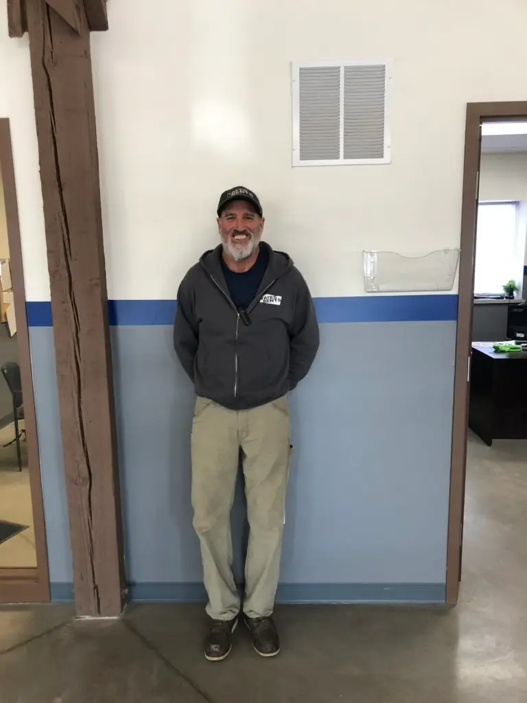 A smiling man with a beard and black baseball cap stands facing forward, wearing a dark gray zippered hoodie with "YATES RENTS" logo, khaki pants, and brown shoes. He stands between a dark wooden beam on the left and a doorway on the right. The wall behind him is white with a blue stripe and light gray lower section, featuring a white rectangular vent and a clear plastic wall pocket.