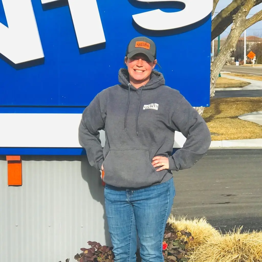 A smiling woman in a gray hoodie and blue jeans stands in front of a blue sign with white lettering. She wears a gray baseball cap with an orange "TATES RENTS" patch. Her hands are on her hips, and she stands next to a low hedge with some red flowers.