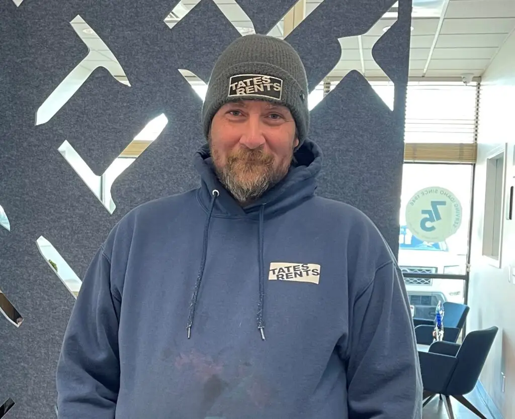 A smiling man with a brown beard and mustache wears a gray beanie with "TATES RENTS" in white text, and a navy blue hooded sweatshirt with a "TATES RENTS" logo on the chest. He stands against a dark gray textured wall with geometric reflective cutouts. In the background, a glass door reveals reflections of outdoors and a blue chair.