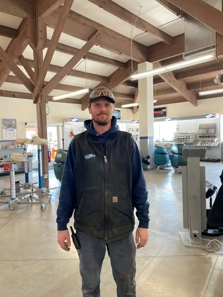 A man with a light beard and short brown hair smiles faintly while standing in a large indoor space. He wears a dark blue hoodie, a black work vest with a "TATES RENTS" logo, grey work pants, and a camouflage baseball cap with clear safety glasses pushed up on it. A walkie-talkie is clipped to his right hip. The background shows exposed wooden ceiling beams, fluorescent lighting, and various rental equipment or store displays, including floor polishers and lifting equipment.