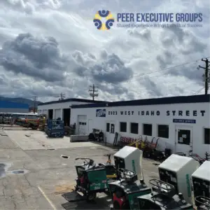 A rental yard with a white building labeled "TATES RENTS" and "2923 WEST IDAHO STREET", featuring several garage doors and windows. Various construction equipment, including ride-on power trowels (some labeled "WHITEMAN") and a blue scissor lift, are parked on the asphalt lot under a cloudy sky.