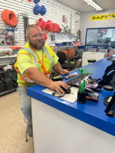 A bearded man with a light-colored beard and short hair, wearing a bright yellow t-shirt and an orange and yellow reflective safety vest, sits at a blue counter. He is smiling slightly and looking at the viewer while operating a black computer mouse on a mousepad. In the background, white slat walls display various hardware items and tools, including coiled orange hoses, red gasoline cans, blue hard hats, and ladders. A yellow sign with "SAFVIA" is visible on the wall.
