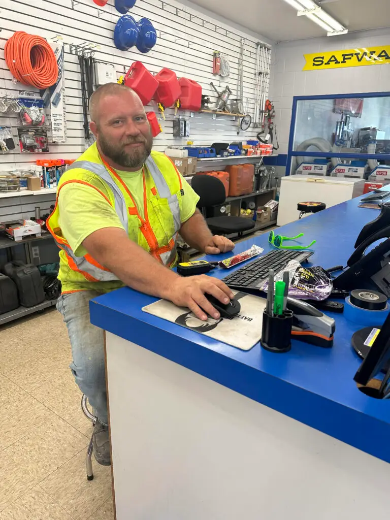 A bearded man with a light-colored beard and short hair, wearing a bright yellow t-shirt and an orange and yellow reflective safety vest, sits at a blue counter. He is smiling slightly and looking at the viewer while operating a black computer mouse on a mousepad. In the background, white slat walls display various hardware items and tools, including coiled orange hoses, red gasoline cans, blue hard hats, and ladders. A yellow sign with "SAFVIA" is visible on the wall.