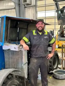A smiling man named David, wearing a grey work shirt with reflective stripes and a "TATES RENTS" logo, stands next to an open blue industrial vehicle in a garage. He has a beard and a baseball cap with sunglasses. A large radiator is visible on the vehicle, and a yellow construction machine is in the background.