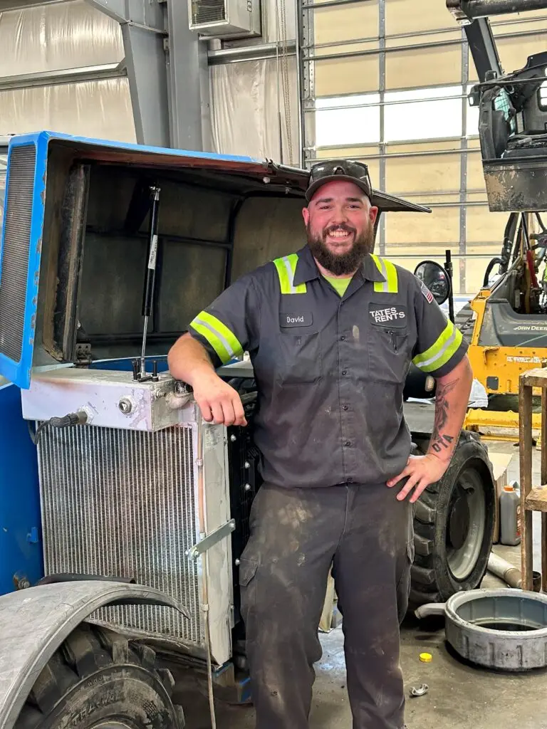 A smiling man named David, wearing a grey work shirt with reflective stripes and a "TATES RENTS" logo, stands next to an open blue industrial vehicle in a garage. He has a beard and a baseball cap with sunglasses. A large radiator is visible on the vehicle, and a yellow construction machine is in the background.