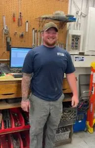 A smiling man with a beard and a full sleeve tattoo on his left arm, wearing a blue t-shirt with "TATES RENTS" logo, grey work pants, and a green baseball cap. He stands in a workshop with a pegboard displaying various tools, a computer monitor, and shelves with parts and equipment in the background.