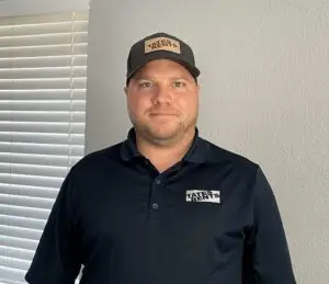 A man with light skin, blue eyes, and a short beard smiles slightly, wearing a dark grey polo shirt with "TATES RENTS" logo on the left chest, and a dark grey baseball cap with a light brown "TATES RENTS" patch. White horizontal blinds are visible on the left side of the frame.