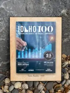 A rectangular plaque featuring the 'Idaho 100' logo on a blue background, with a hand interacting with a digital bar and line graph showing growth. Text reads 'SPECIAL ADVERTISING FEATURE | NOVEMBER 2023', 'RECOGNIZING IDAHO'S TOP PRIVATE COMPANIES', 'Representing over $55 billion in Idaho business revenue', and 'ON THE RISE 71% of Idaho 100 companies experience revenue growth'. A circular logo celebrates '25 YEARS PRIVATE COMPANIES'. Logos for Idaho Trust Bank, KPMG, Holland & Hart, and Idaho Statesman are visible. At the bottom, 'Tates Rents - #89' is printed. The plaque rests on dark grey stone and light-colored river pebbles with dried leaves.