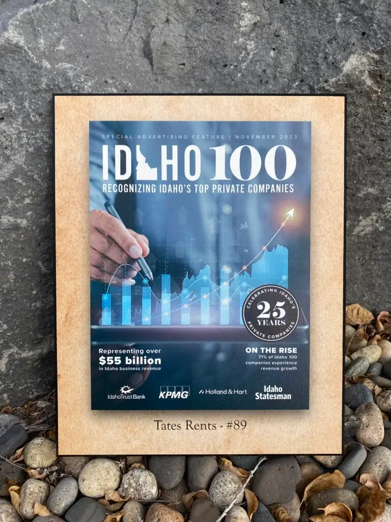A rectangular plaque featuring the 'Idaho 100' logo on a blue background, with a hand interacting with a digital bar and line graph showing growth. Text reads 'SPECIAL ADVERTISING FEATURE | NOVEMBER 2023', 'RECOGNIZING IDAHO'S TOP PRIVATE COMPANIES', 'Representing over $55 billion in Idaho business revenue', and 'ON THE RISE 71% of Idaho 100 companies experience revenue growth'. A circular logo celebrates '25 YEARS PRIVATE COMPANIES'. Logos for Idaho Trust Bank, KPMG, Holland & Hart, and Idaho Statesman are visible. At the bottom, 'Tates Rents - #89' is printed. The plaque rests on dark grey stone and light-colored river pebbles with dried leaves.