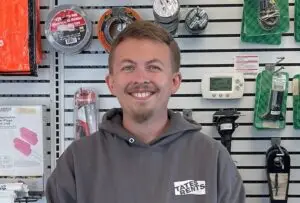 A smiling man with short brown hair and a goatee stands in front of a white pegboard wall displaying various hardware items. He is wearing a grey hooded sweatshirt with "TATES RENTS" in white lettering on the chest. Visible items on the wall include rolls of tape, packaged earplugs, and a white digital thermostat.