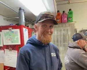 A smiling man with a reddish-blond beard and gray baseball cap wearing sunglasses, and a blue hoodie with a "TATES RENT" logo. Behind him, a red cabinet with papers, a grey water bottle, and three colorful plastic bottles are visible. Another man in a grey hoodie is partially visible on the right.
