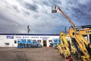 A white rental store building with "TATES RENTS 2923 WEST IDAHO STREET" in blue lettering. Several blue scissor lifts are parked in front. On the right, yellow excavators and a red and yellow boom lift with its arm extended upwards are visible under a cloudy sky.