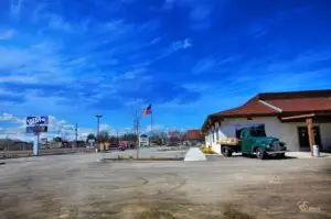 A green vintage flatbed truck parked outside a light-colored building with a brown roof under a bright blue sky with wispy clouds. In the background, a "TATES RENTS" sign is visible, along with other businesses and a US flag flying.