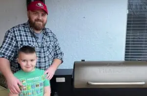A man with a beard and a red 49ers cap stands next to a smiling young boy in a green Minecraft t-shirt. They are beside a dark gray and bronze Traeger pellet grill, with a white textured wall in the background.