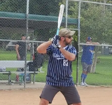 A man in a blue and white pinstriped softball jersey, gray shorts, and a tan cap holds a white and light blue bat over his right shoulder, ready to swing. He wears sunglasses and batting gloves. A chain link fence is in the background, with other players visible behind it.