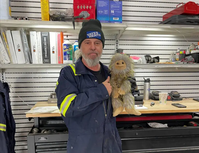 A man with a gray beard and a black "TATES RENTS" beanie smiles while holding a furry, light brown toy and giving a thumbs-up. He wears a dark blue work jumpsuit with reflective yellow stripes. In the background, there are white shelves stocked with binders labeled "Ditch Witch," "NEW HOLLAND," and "BRUSH CHIPPERS Resource Library," along with various tools and fluids like "NAPA Brake Fluid."