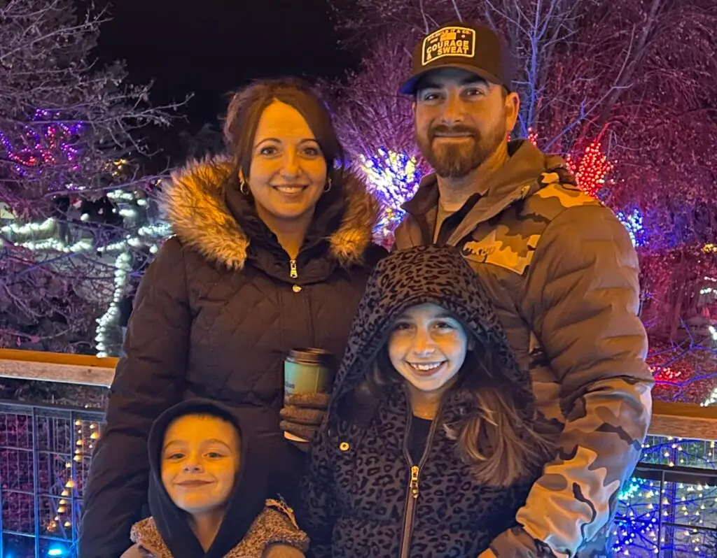 A smiling family of four, two adults and two children, stands outdoors at night with a background of trees adorned with colorful string lights. The woman wears a black fur-lined puffer coat and holds a coffee cup, the man wears a camouflage puffer jacket and a black baseball cap with "COURAGE & SWEAT" text, the girl wears a leopard print hooded coat, and the boy wears a hooded jacket.