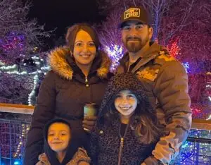 A smiling family of four, two adults and two children, stands outdoors at night with a background of trees adorned with colorful string lights. The woman wears a black fur-lined puffer coat and holds a coffee cup, the man wears a camouflage puffer jacket and a black baseball cap with "COURAGE & SWEAT" text, the girl wears a leopard print hooded coat, and the boy wears a hooded jacket.
