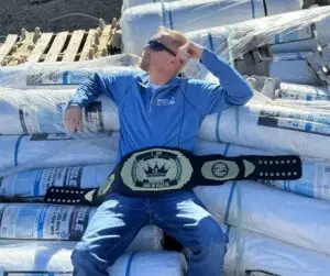 A man with a beard and sunglasses wearing a blue shirt and jeans lies on a stack of white plastic sheeting rolls, holding a black and gold championship belt with "I AM THE UNDISPUTED KING OF PLASTIC" across his waist. The rolls are labeled as "REINFORCED POLY" and "FLAME RETARDANT".