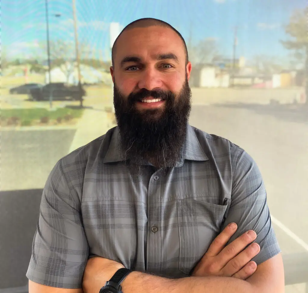 Bald man with a dark beard smiling, wearing a gray plaid short-sleesleeved shirt with arms crossed, in front of a blurred outdoor background.