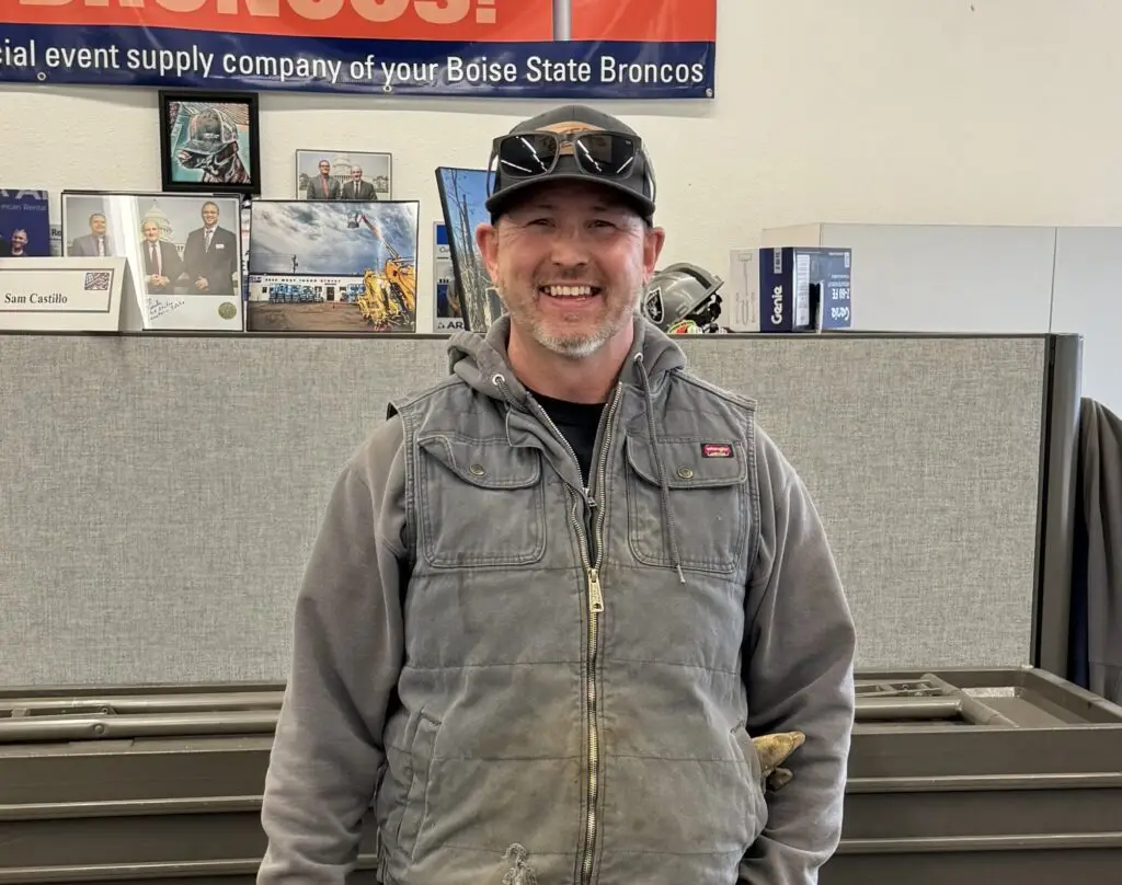 A smiling man with a beard and baseball cap, wearing sunglasses on his cap, a gray hooded sweatshirt, and a gray puffer vest with a "Wrangler" patch. He stands in front of a gray cubicle wall with framed pictures and a "Boise State Broncos" banner in the background.f
