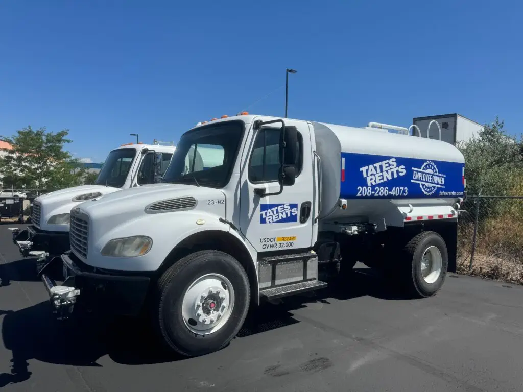 A white Tates Rents water truck, marked "23-02", with a large cylindrical tank displaying "TATES RENTS", "208-286-4073", "EMPLOYEE OWNED", and "tatesrents.com". It has "USDOT 937401 GVWR 32,000" on the side, parked on asphalt under a clear blue sky, with parts of another identical truck visible in the background and a chain-link fence on the right.