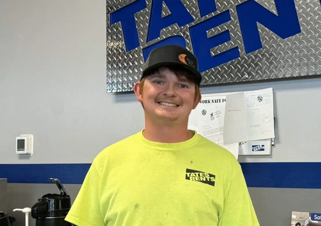 A smiling young man with reddish-brown hair and a light mustache, wearing a black baseball cap with a "Ditch Witch" logo and a bright yellow long-sleeved shirt with a "TATES RENTS" logo. He stands in front of a gray wall with a blue stripe, and a large blue sign with silver diamond plate texture showing part of the word "TATEN" is visible behind him.