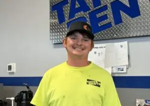 A smiling young man with reddish-brown hair and a light mustache, wearing a black baseball cap with a "Ditch Witch" logo and a bright yellow long-sleeved shirt with a "TATES RENTS" logo. He stands in front of a gray wall with a blue stripe, and a large blue sign with silver diamond plate texture showing part of the word "TATEN" is visible behind him.