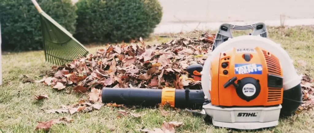 An orange and white Stihl BR 700 leaf blower with a black nozzle attachment and a blue "Tate's Rents" sticker sits on green grass next to a large pile of brown autumn leaves. A green rake leans into the leaf pile in the background, with green bushes beyond.