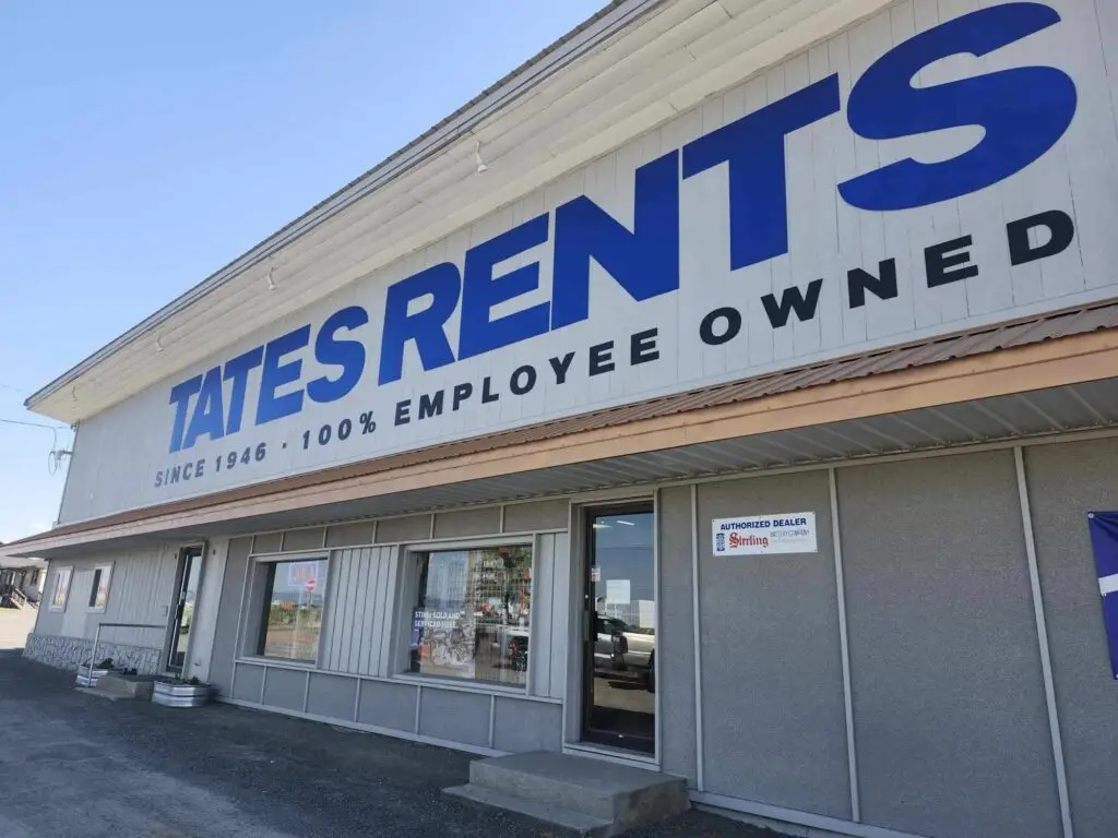 A white and grey commercial building with a large blue sign reading "TATES RENTS" and "SINCE 1946 - 100% EMPLOYEE OWNED". The building has multiple windows and a black door, with a blue banner displaying "TATES RENTS" hanging on the grey section. The foreground is a dark gravel lot, and in the background, there is a clear blue sky, green grass, trees, and distant buildings.