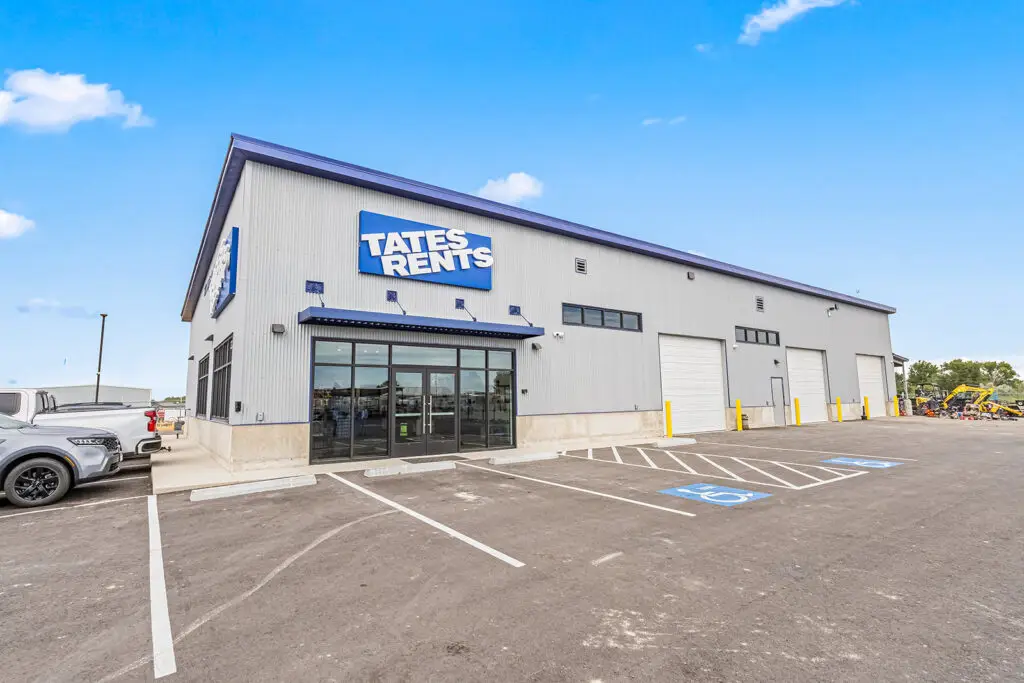 A modern industrial building with gray corrugated metal siding and a blue roof trim, featuring a prominent "TATES RENTS" sign in blue. The building has large glass double doors at the entrance, multiple horizontal windows, and three white roll-up garage doors. A paved parking lot with white lines and two blue handicap parking symbols is in the foreground. Several vehicles, including a white pickup truck and a gray SUV, are parked to the left, and heavy equipment is visible in the background on the right.