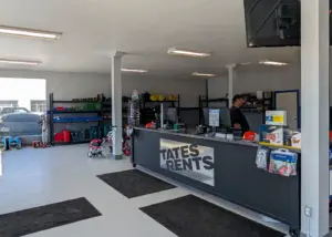 Interior view of a rental shop with a black counter displaying "TATES RENTS" in silver diamond plate. A man in a black shirt and cap stands behind the counter. Shelves along the back wall hold various tools and equipment like red generators, blue and green objects, and yellow bins. The floor is light gray with several black floor mats. Overhead, fluorescent lights illuminate the space. A large window on the left shows a parked vehicle.