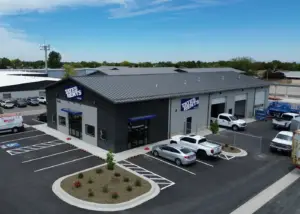 An aerial view of a Tates Rents commercial building with dark gray corrugated metal and light gray walls under a blue sky. The building has multiple garage bays, some open with white pickup trucks visible. A paved parking lot with white stripes includes a handicap spot and several parked vehicles, a silver car, a white pickup, and a white delivery van. Landscaped islands with small plants and shrubs are in the foreground.