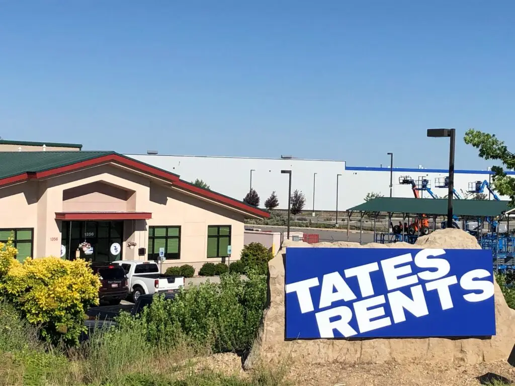 A commercial building with a beige stucco exterior and a dark green sloped roof. In the foreground, a tall black sign displays "TATE'S RENTS" in white letters on a blue background. Multiple cars, including a black Mini Cooper with a red and black circular logo on the hood, are parked in the lot in front of the building. Green bushes and a tree partially obscure the view on the left and right.