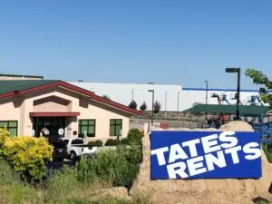 A commercial building with a beige stucco exterior and a dark green sloped roof. In the foreground, a tall black sign displays "TATE'S RENTS" in white letters on a blue background. Multiple cars, including a black Mini Cooper with a red and black circular logo on the hood, are parked in the lot in front of the building. Green bushes and a tree partially obscure the view on the left and right.