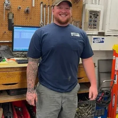 A smiling man with a beard and a full sleeve tattoo on his left arm, wearing a blue t-shirt with "TATES RENTS" logo, grey work pants, and a green baseball cap. He stands in a workshop with a pegboard displaying various tools, a computer monitor, and shelves with parts and equipment in the background.